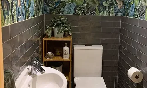 Stylish small powder room with dark subway tiles, floral wallpaper, and natural wood shelving designed by Bathrooms Renovations Hamilton.